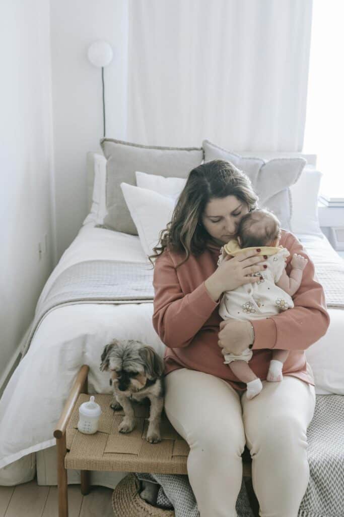 Happy mother sitting on chair at bed with puppy and hugging small baby inn light bedroom