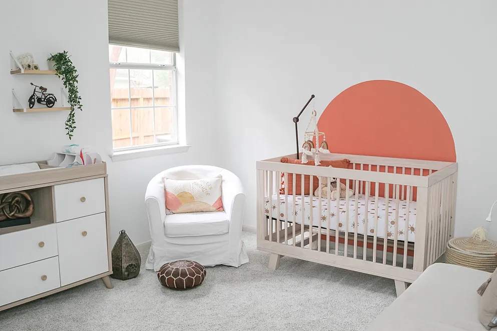 A minimalist baby nursery features a light wood crib, a terracotta sun mural, a matching changing table, and a white armchair